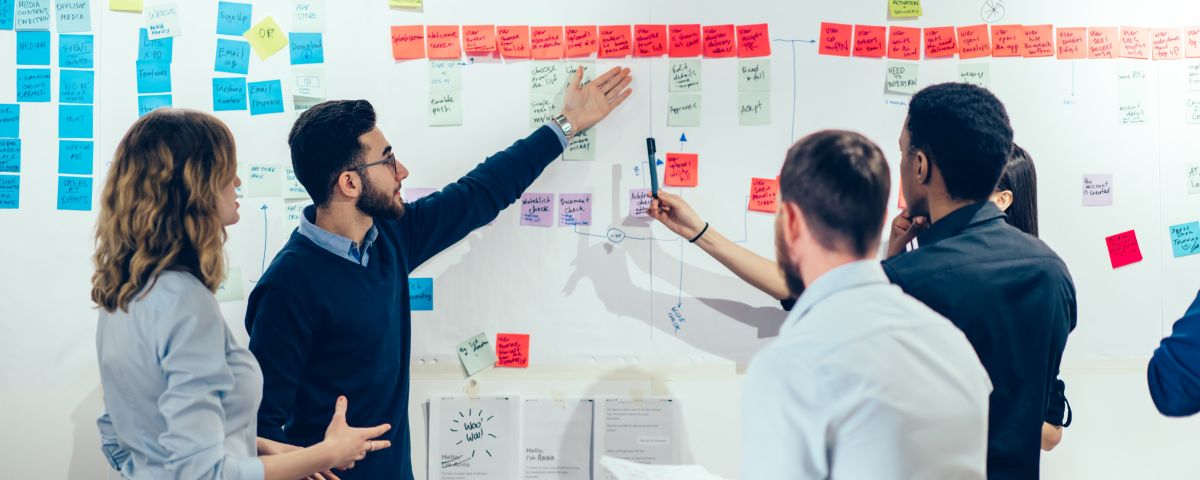 Intelligent male professional pointing on colorful stickers with text message glueded on wall and discussing information with creative multicultural colleagues having brainstorming meeting in office Team diskutiert Prozessablauf als Symbol für Service- und Prozessinnovation - TOM SPIKE