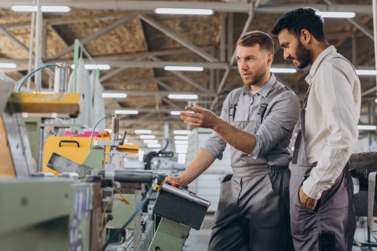 A male inspector or operator of a workshop for the production of aluminum and plastic wreaths trains an intern. International team of men working together near a machine in a factory.