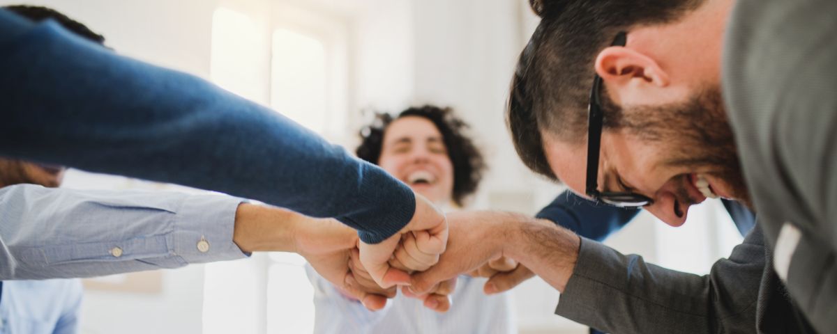 Young businesspeople sitting around table in a modern office, making fist bump. Innovationsmanagerin und Partner stecken die Fäuste zusammen und lachen - TOM SPIKE