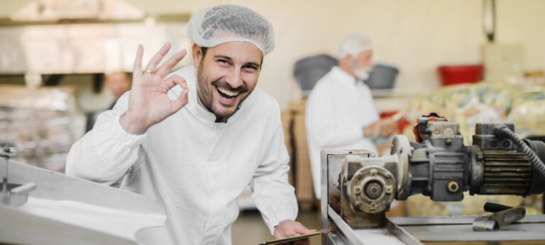 lachender Produktionsmitarbeiter mit Haarnetz an rustikaler Maschine als Symbol für Innovation in der Produktion - TOM SPIKE